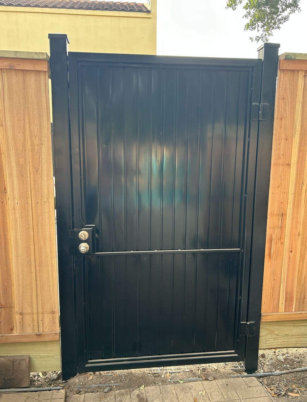 Black metal gate with vertical slats and a locking mechanism beside a wooden fence.