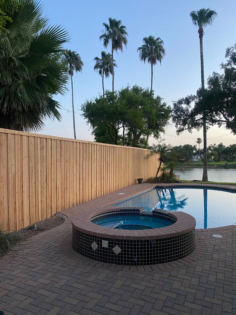 Peaceful backyard with a pool and hot tub, surrounded by palm trees and a wooden fence.