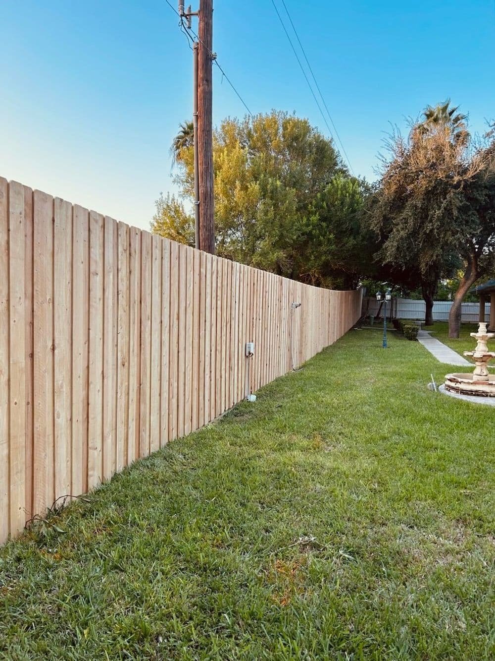 Wooden fence extending along a green lawn with trees and a fountain in the background.