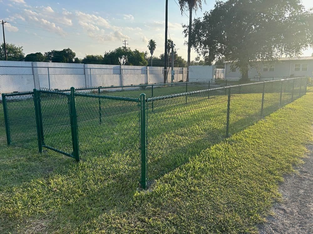 Green chain-link fence enclosing a grassy area with trees under a clear blue sky.