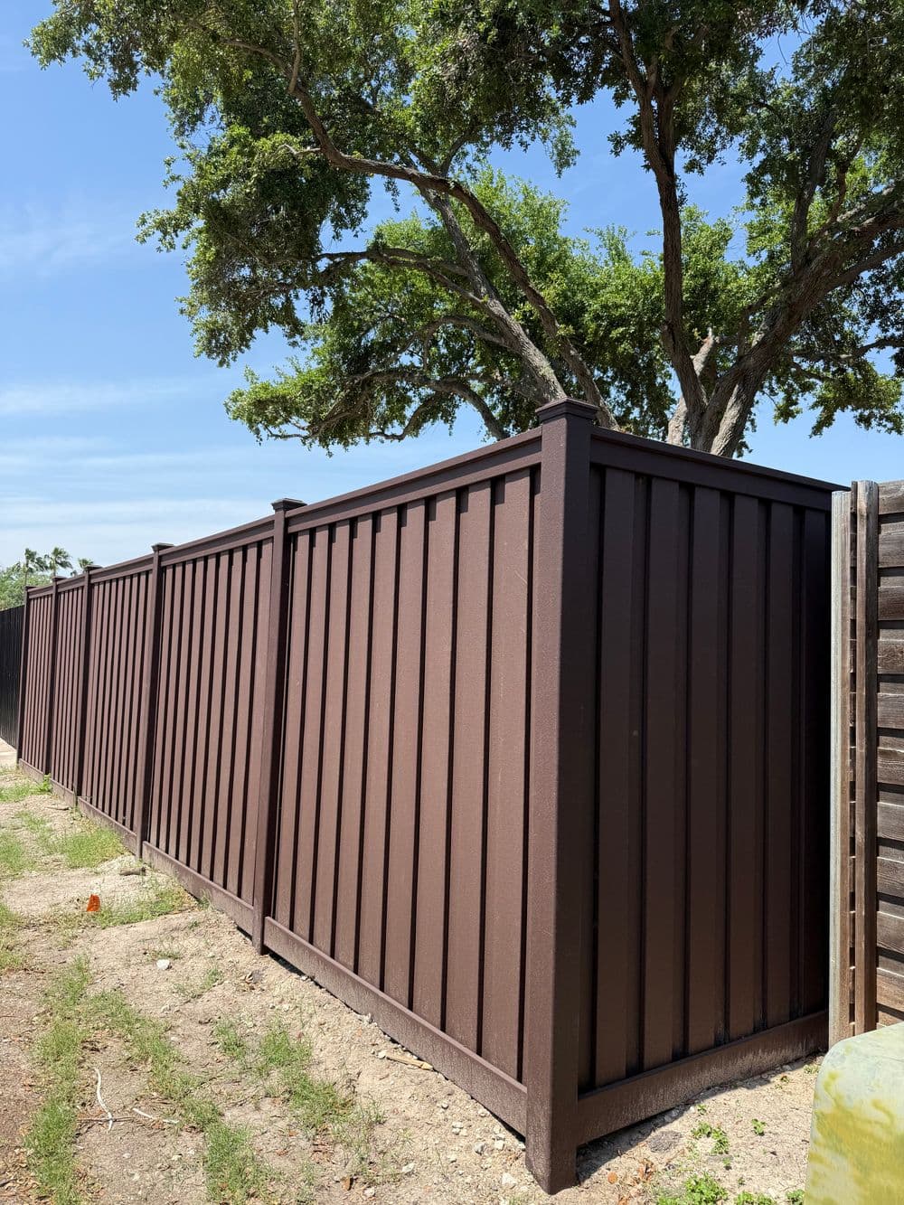 Brown wooden fence along a grassy area with large trees and clear blue sky in the background.